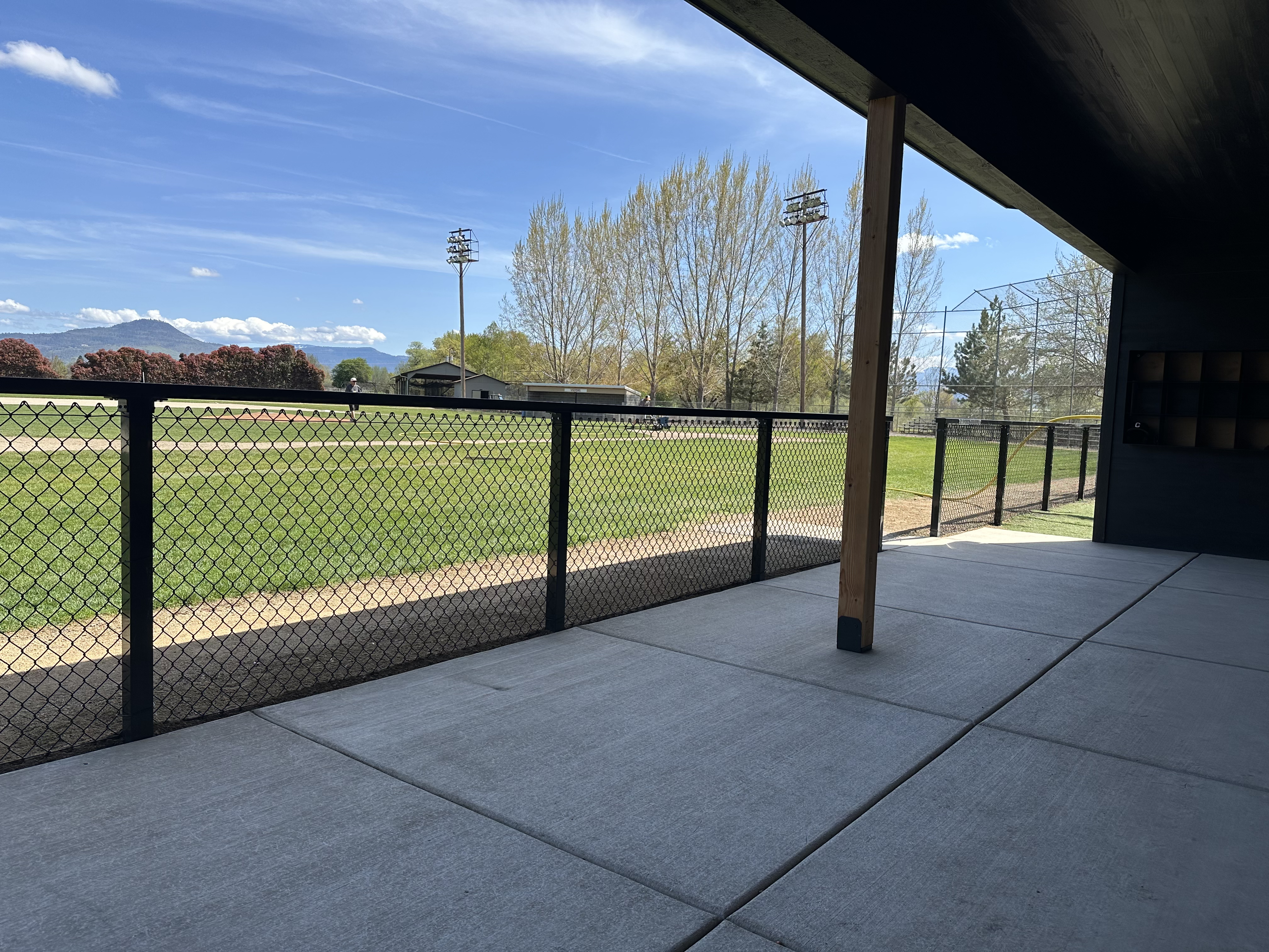 The image shows a covered concrete patio overlooking a grassy baseball field enclosed by a black chain-link fence under a blue sky.