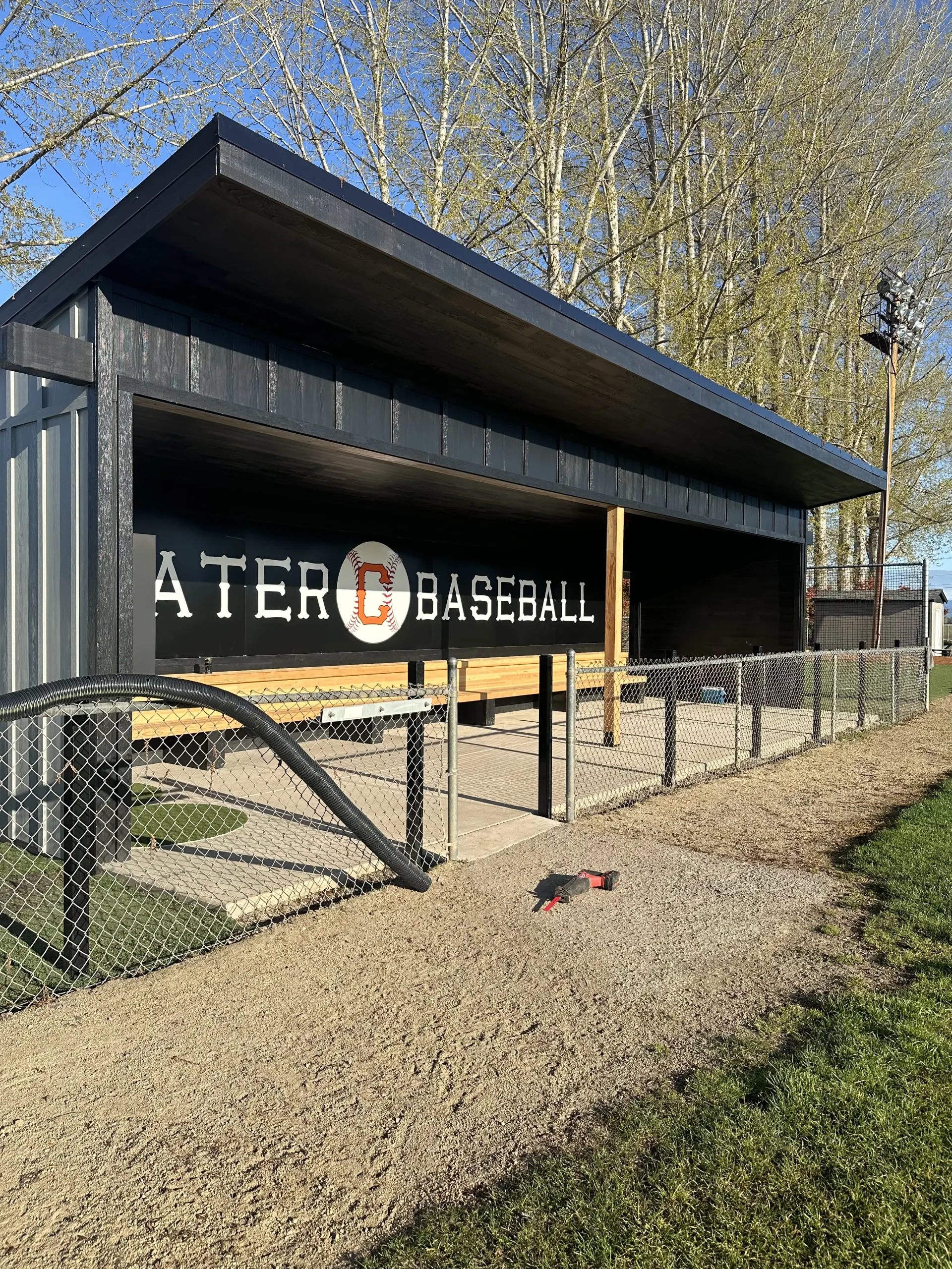 The image shows a newly built or updated modern baseball dugout with a black metal roof, wooden benches, and chain-link fencing.