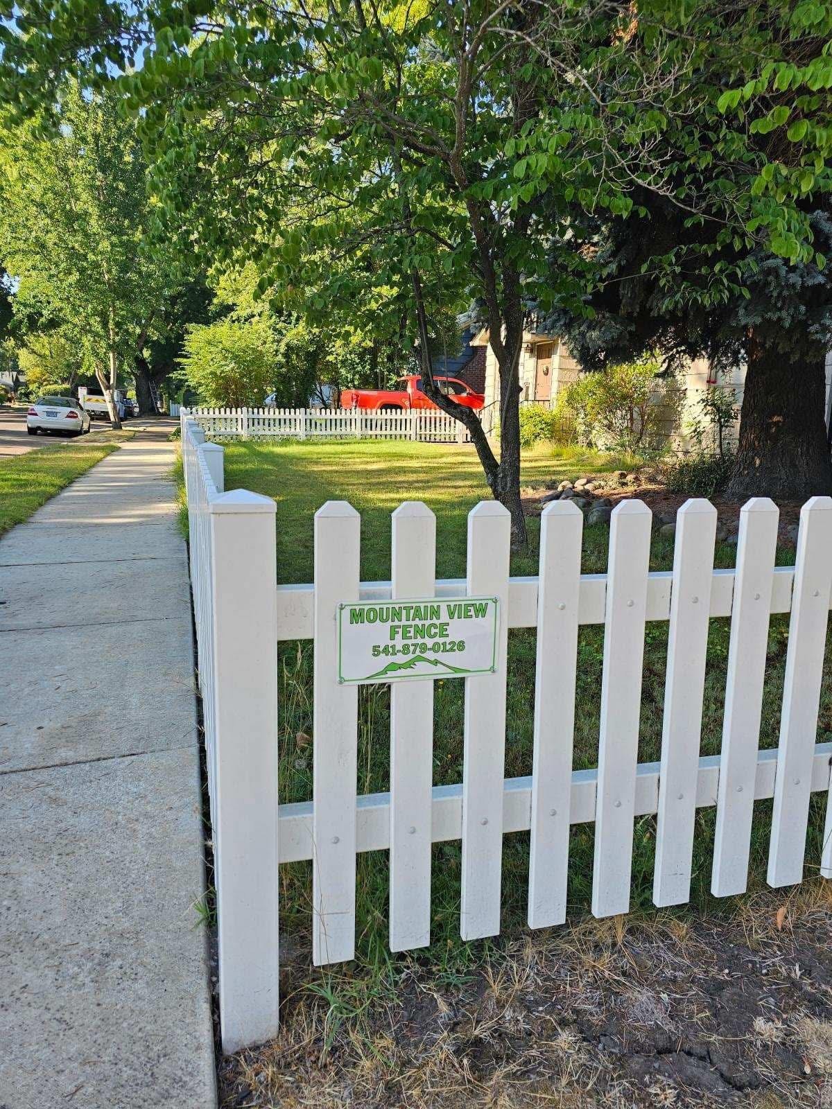 The image shows a white vinyl picket fence with a sign for Mountain View Fence (541-879-0128