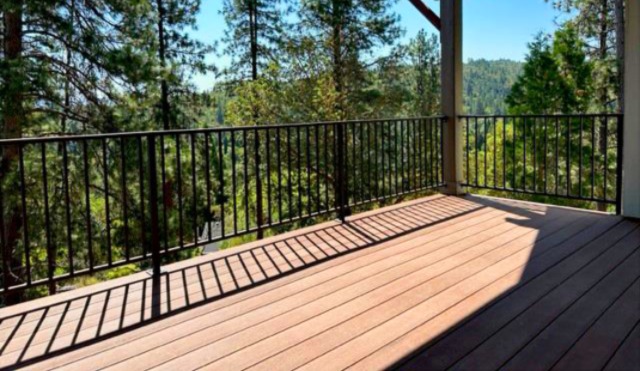 The image shows an outdoor wooden deck with a black metal baluster railing overlooking a green, tree-filled forest